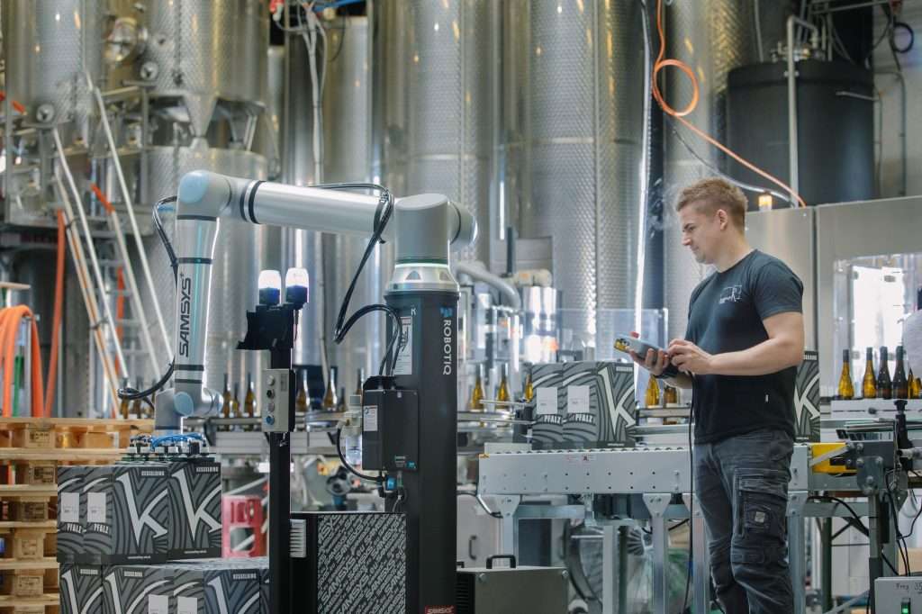 Cobot su colonna di pallettizzazione al lavoro a fine linea in una cantina, con l'operatore accanto